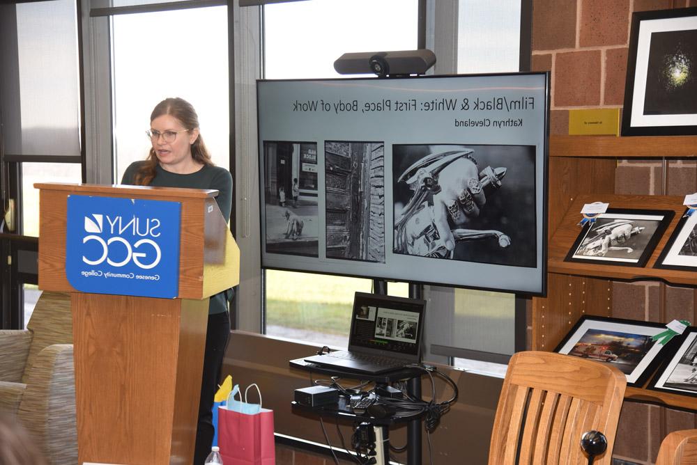 Systems & Electronic Services Librarian Liz Simmons speaking at ceremony, slides showing Film/B&W First Place winning photos behind