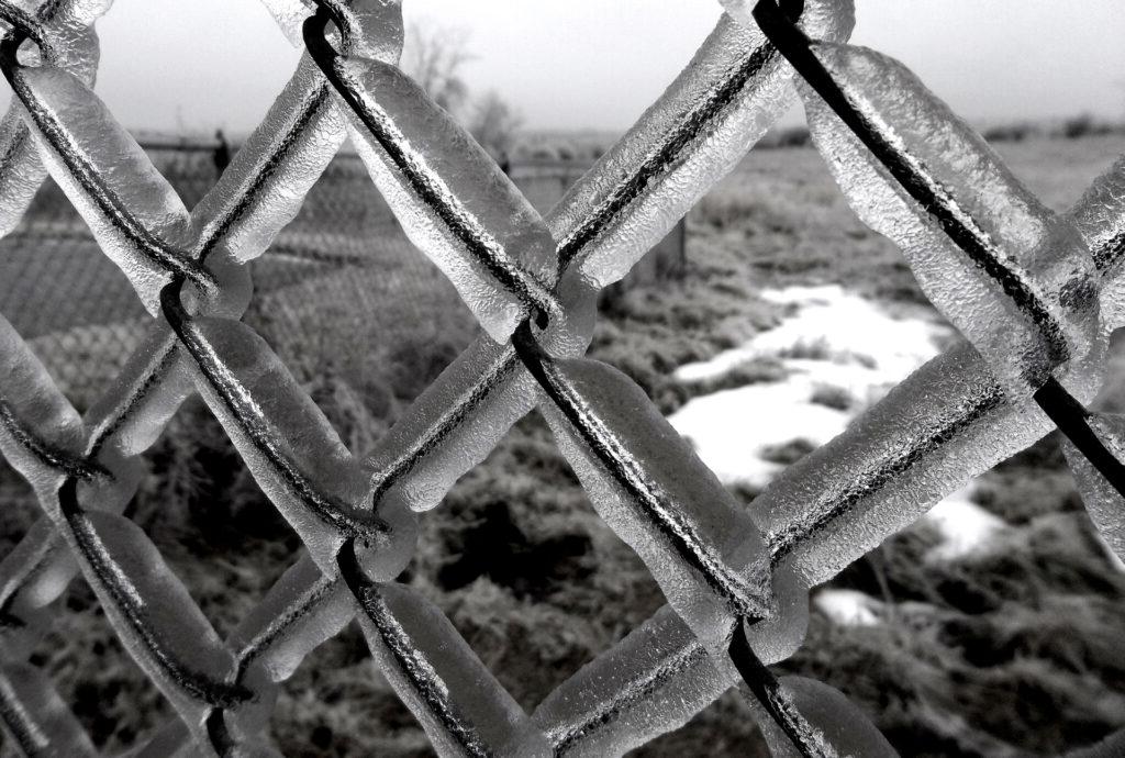 Black and white photo of ice covered fence with patchy snow in background